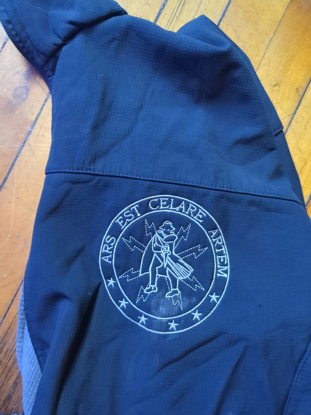 Embroidered roundel reading ARS EST CELARE ARTEM around a trench-coated figure with lightning bolts